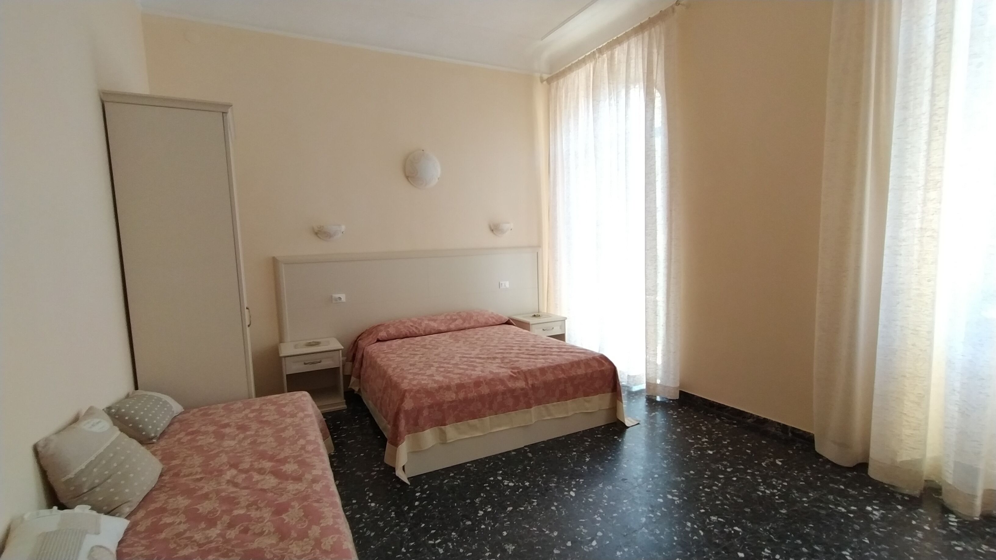 A hotel room with a bed, a bedside table, a nightstand, a wardrobe, and a window with curtains.