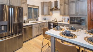 A modern kitchen with stainless steel appliances, a large island with seating, and a view of the outdoors. - Lush Ski-in/out Home! Wood Fireplace, Grill, Walk to Resort! (Big Sky)