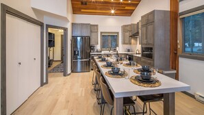 A modern kitchen with a central island, stainless steel appliances, and a view of the outdoors. - Lush Ski-in/out Home! Wood Fireplace, Grill, Walk to Resort! (Big Sky)