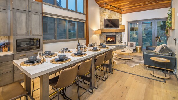 A modern kitchen with a long dining table set for six, a stone fireplace, and a cozy seating area with a leather sofa and a small round table. - Lush Ski-in/out Home! Wood Fireplace, Grill, Walk to Resort! (Big Sky)