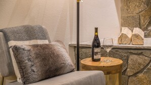 A cozy seating area with a plush chair, a small wooden table with a wine bottle and glass, and a stone wall in the background. - Lush Ski-in/out Home! Wood Fireplace, Grill, Walk to Resort! (Big Sky)