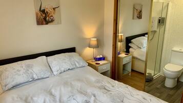 A hotel room with a bed, a nightstand, a lamp, a mirror, a bathroom with a toilet, and a shower.