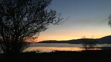 A serene lakeside view at sunset with silhouettes of trees and distant hills.