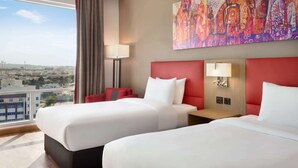 A hotel room with two beds, a red headboard, a large abstract painting, a view of the city, and a balcony with curtains.