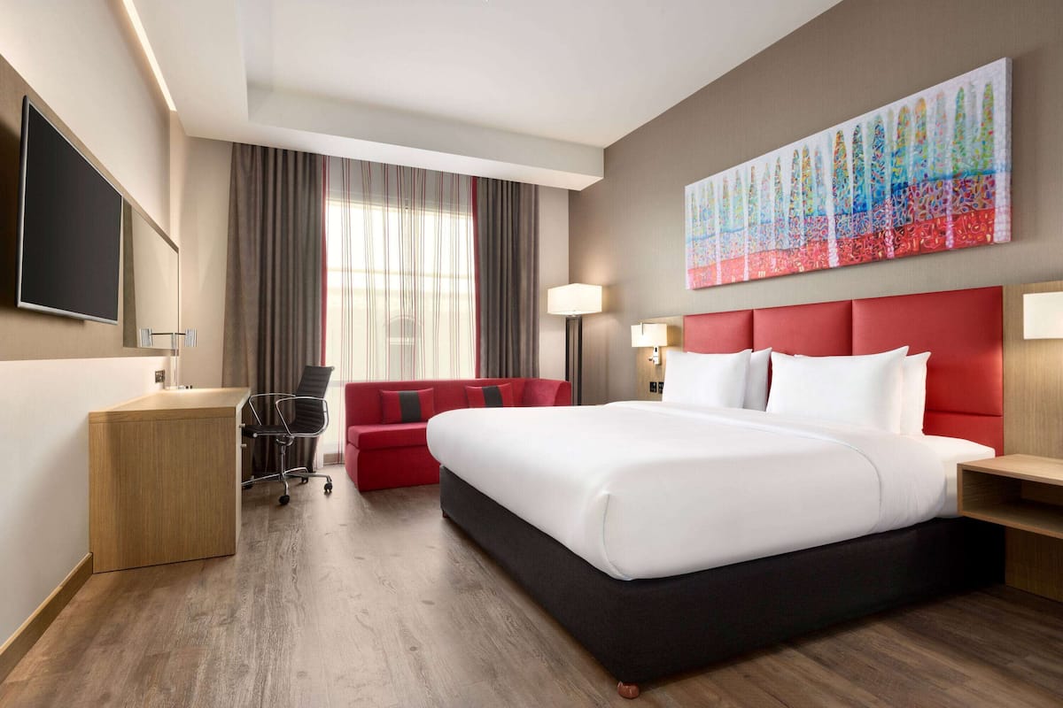 A hotel room with a large bed, a desk, a red sofa, and a large window with curtains.
