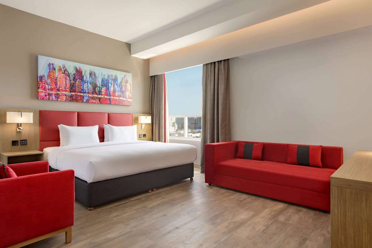 A hotel room with a large bed, a red sofa, a red armchair, a painting on the wall, and a city view through the window.