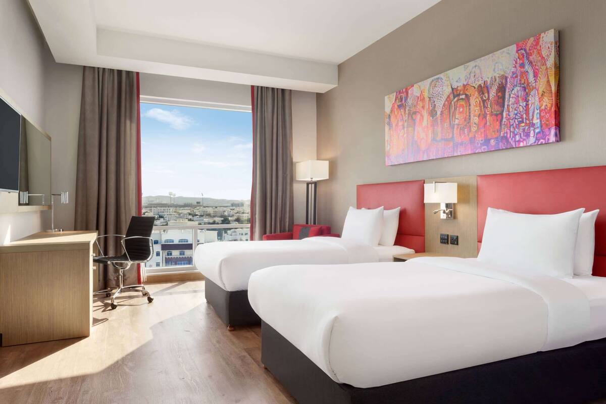 A hotel room with two beds, a desk, a chair, and a large window offering a city view.