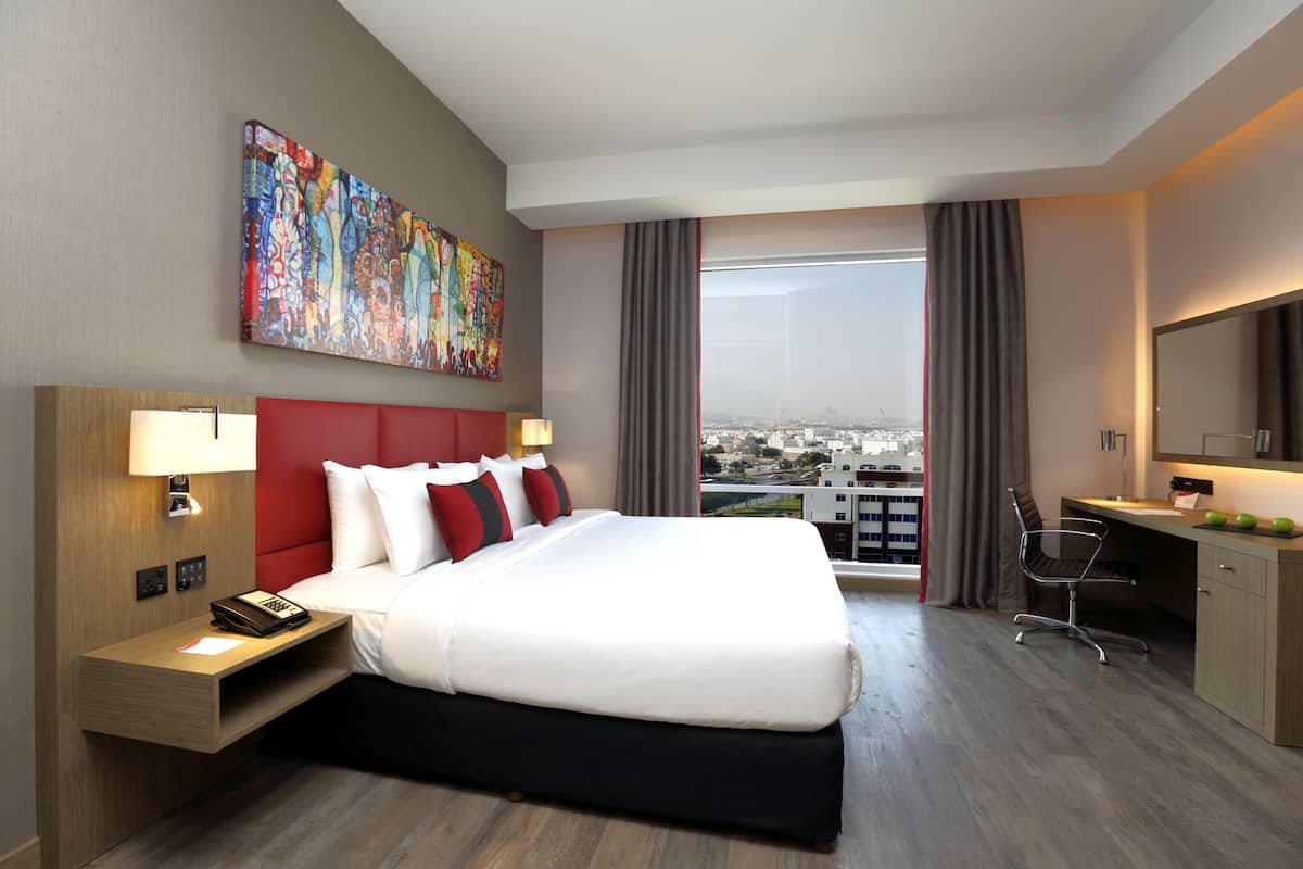 A hotel room with a large bed, a desk, a chair, and a city view.