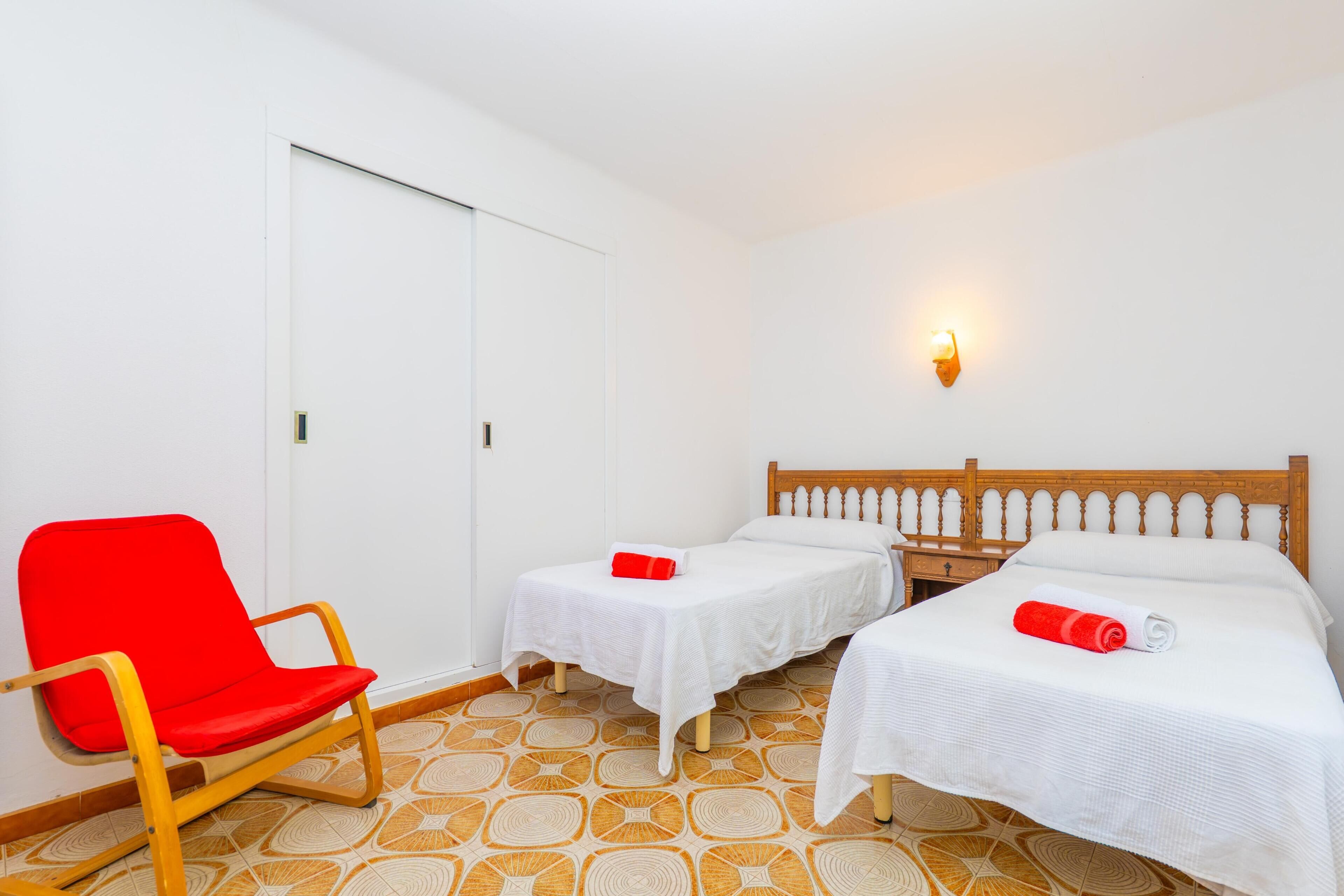 A hotel room with two single beds, a red chair, and a wall-mounted light fixture.