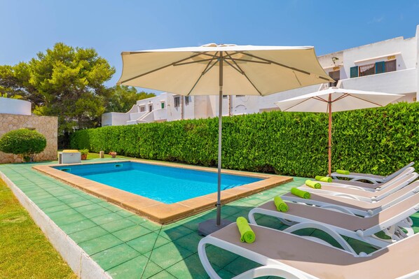 A swimming pool with a sun umbrella, lounge chairs, and a hedge.