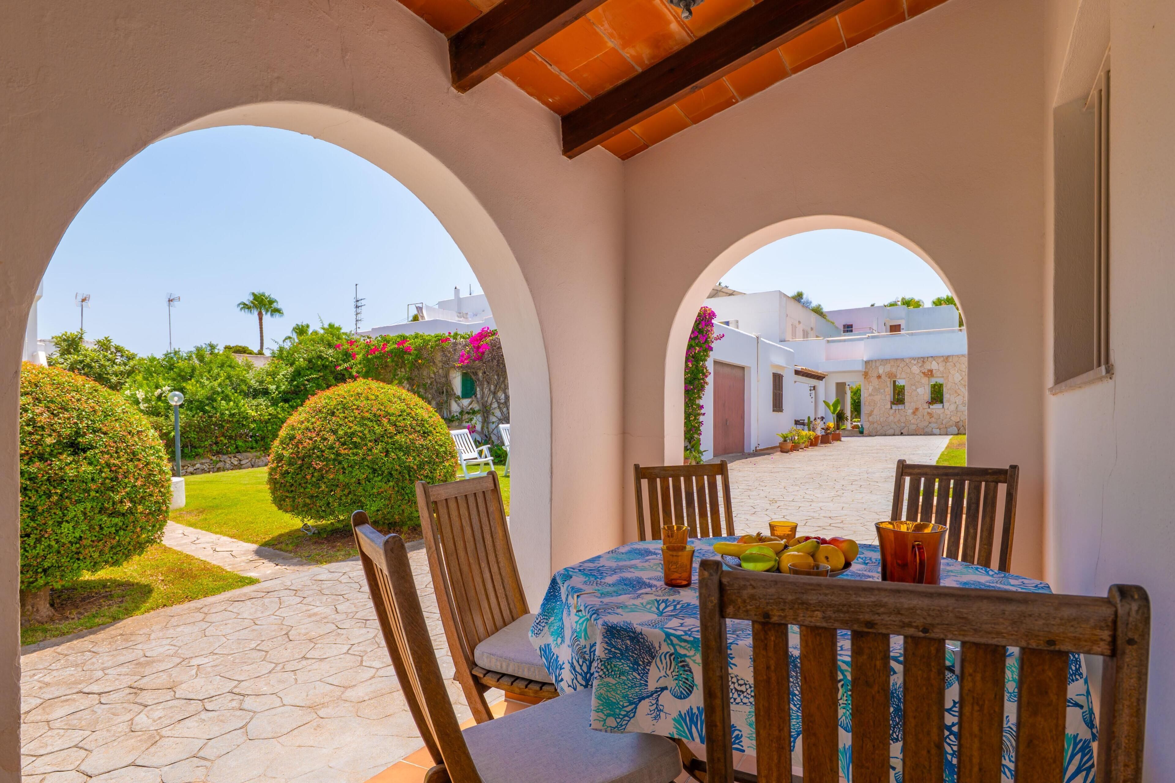 A covered outdoor seating area with a table set for two, surrounded by arches, and a view of a courtyard with neatly trimmed bushes and a pathway.