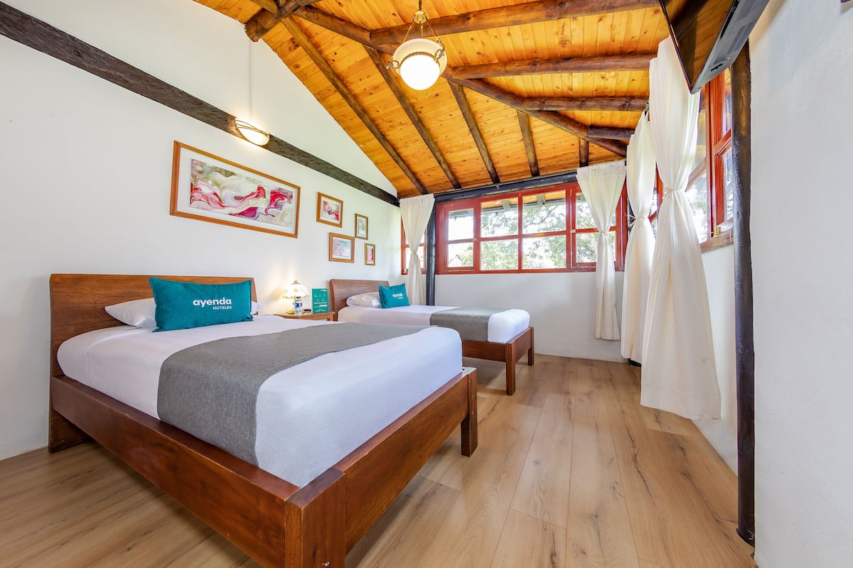 A hotel room with a wooden ceiling, two beds, a nightstand, and a window with curtains.