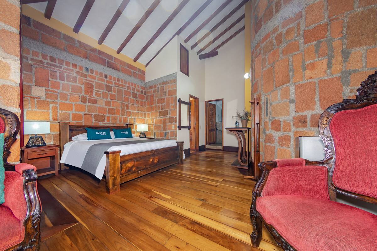 A bedroom with a brick wall, wooden floor, a bed with a white and gray bedspread, two bedside tables with lamps, and a red upholstered chair.