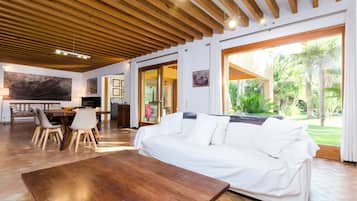 Amplia sala de estar con techo de madera, un sofá blanco, una mesa de comedor con sillas y vista a un jardín a través de grandes ventanales.