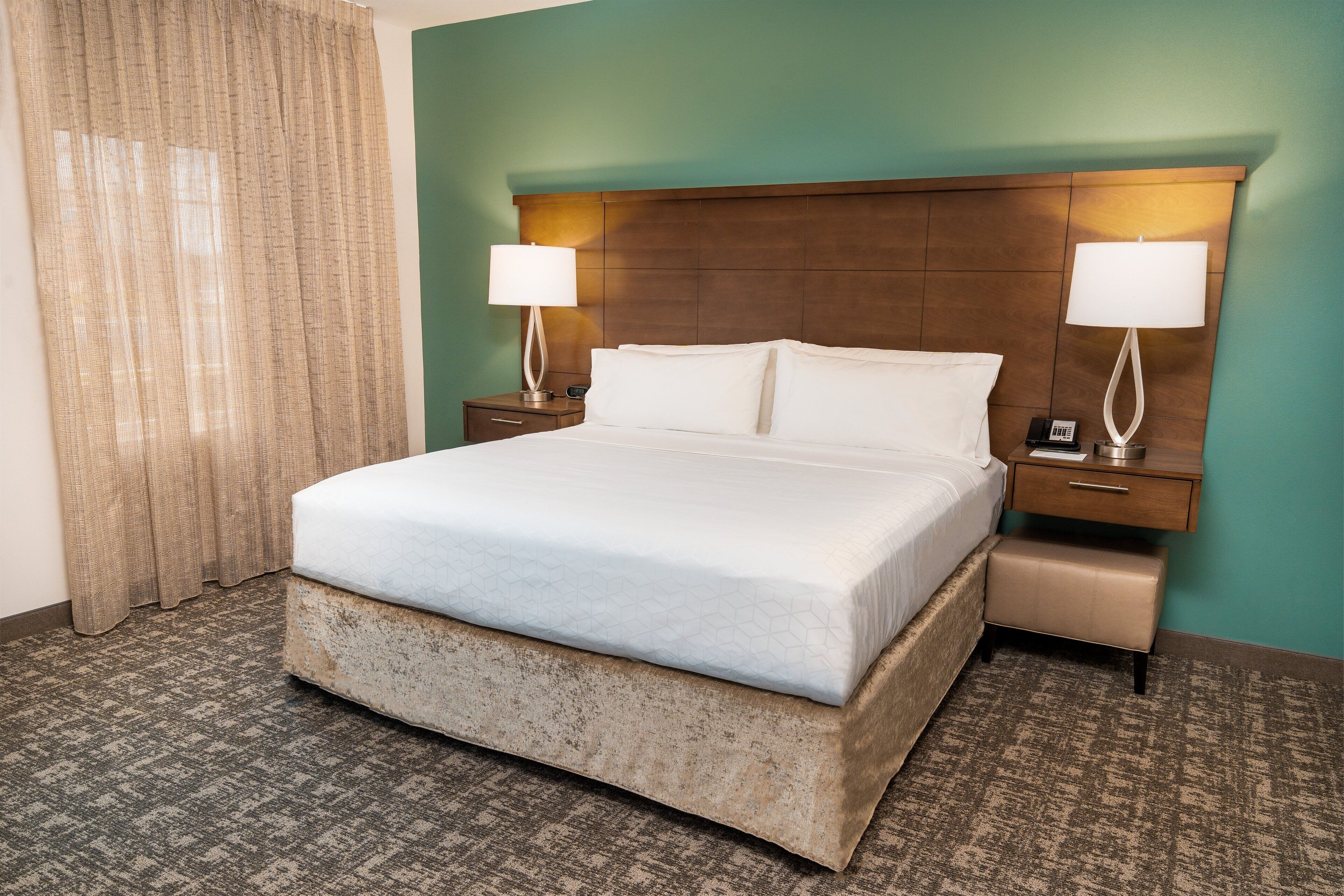 A hotel room with a large bed, two bedside tables, a nightstand, a bench, and a curtain.