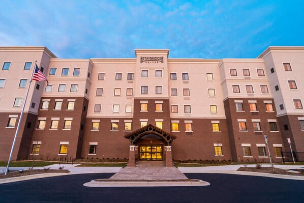 Um hotel de vários andares com entrada central, uma bandeira e uma placa com a inscrição 'Staybridge Suites'.