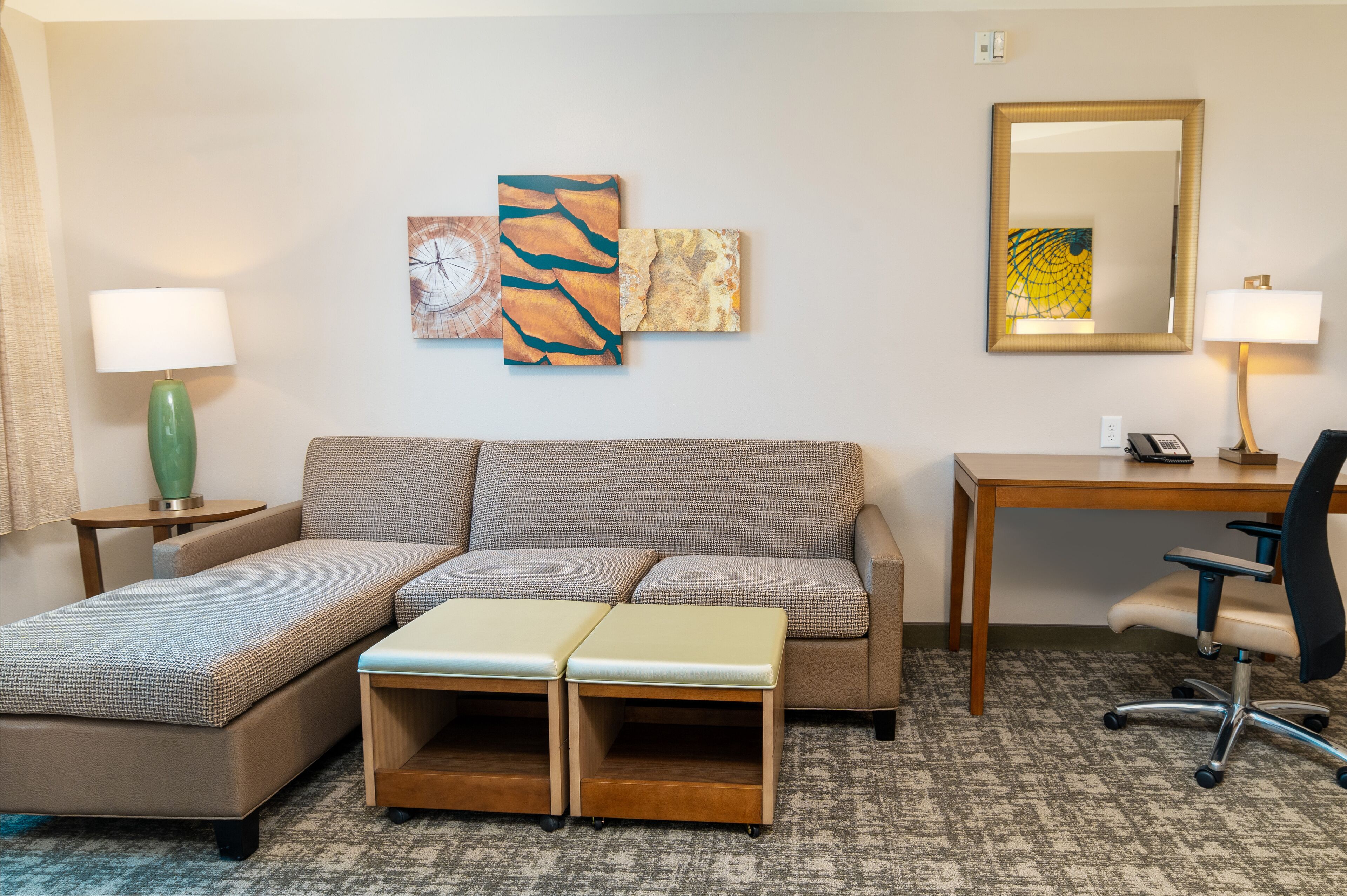 A hotel room with a sofa, a desk, a chair, a lamp, and artwork on the wall.