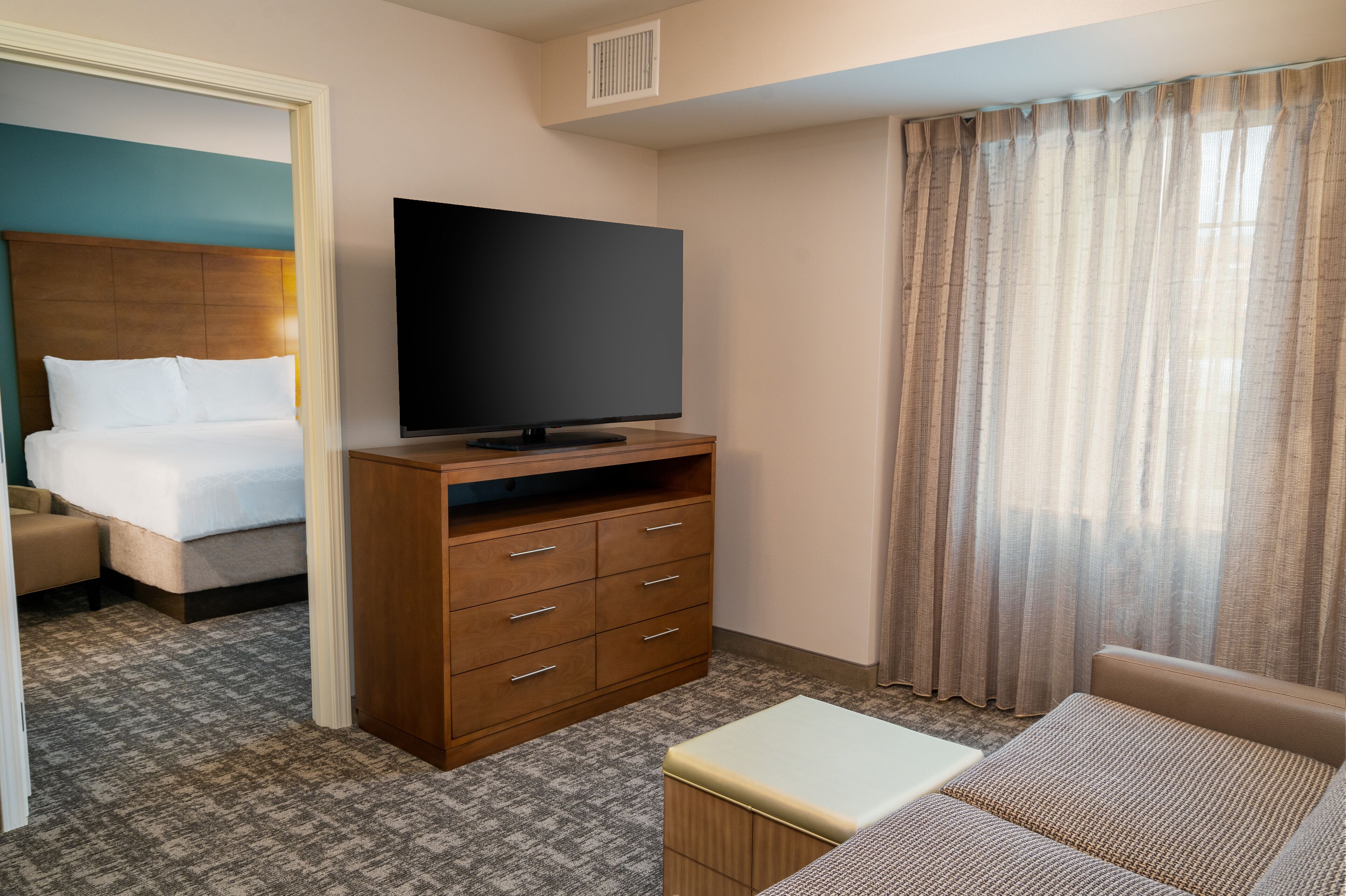 Habitación de hotel con cama, televisión sobre un mueble de madera, sofá y ventana con cortinas.