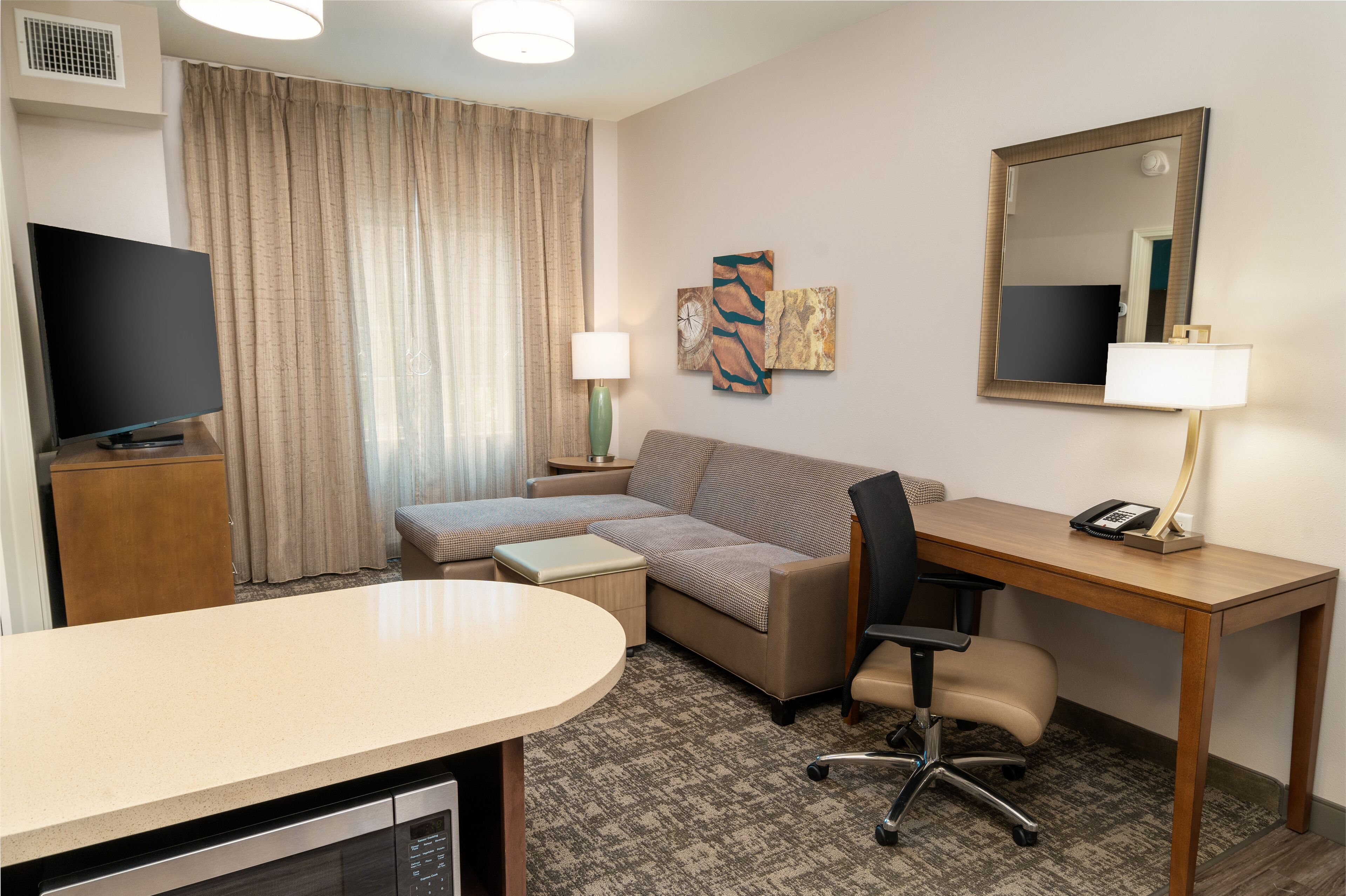 A hotel room with a flat-screen TV, a sofa, a desk, a chair, a lamp, and artwork on the wall.
