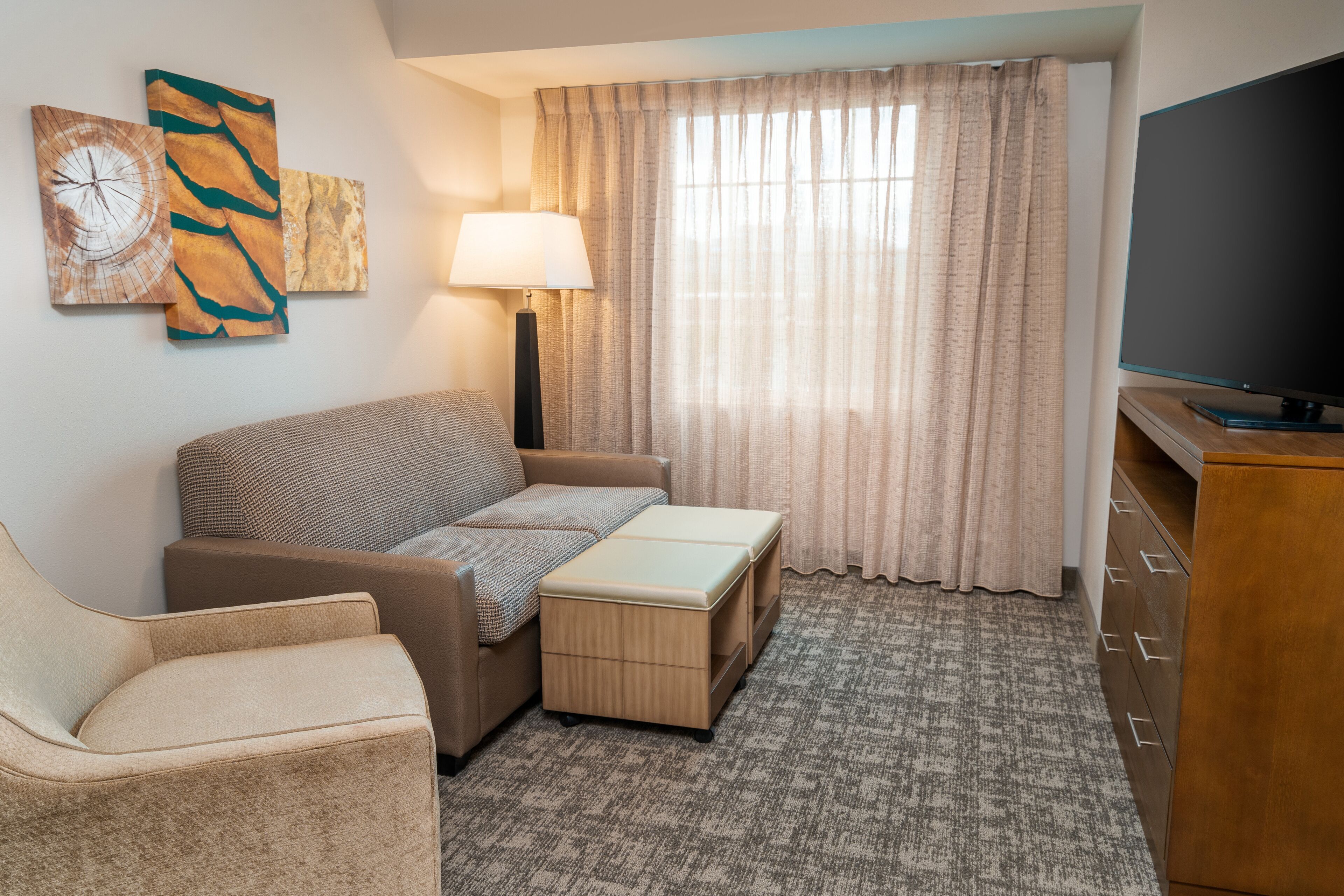 Hotelkamer met een bank, fauteuil, pouf, flatscreen televisie, lamp, gordijnen en kunst aan de muur.
