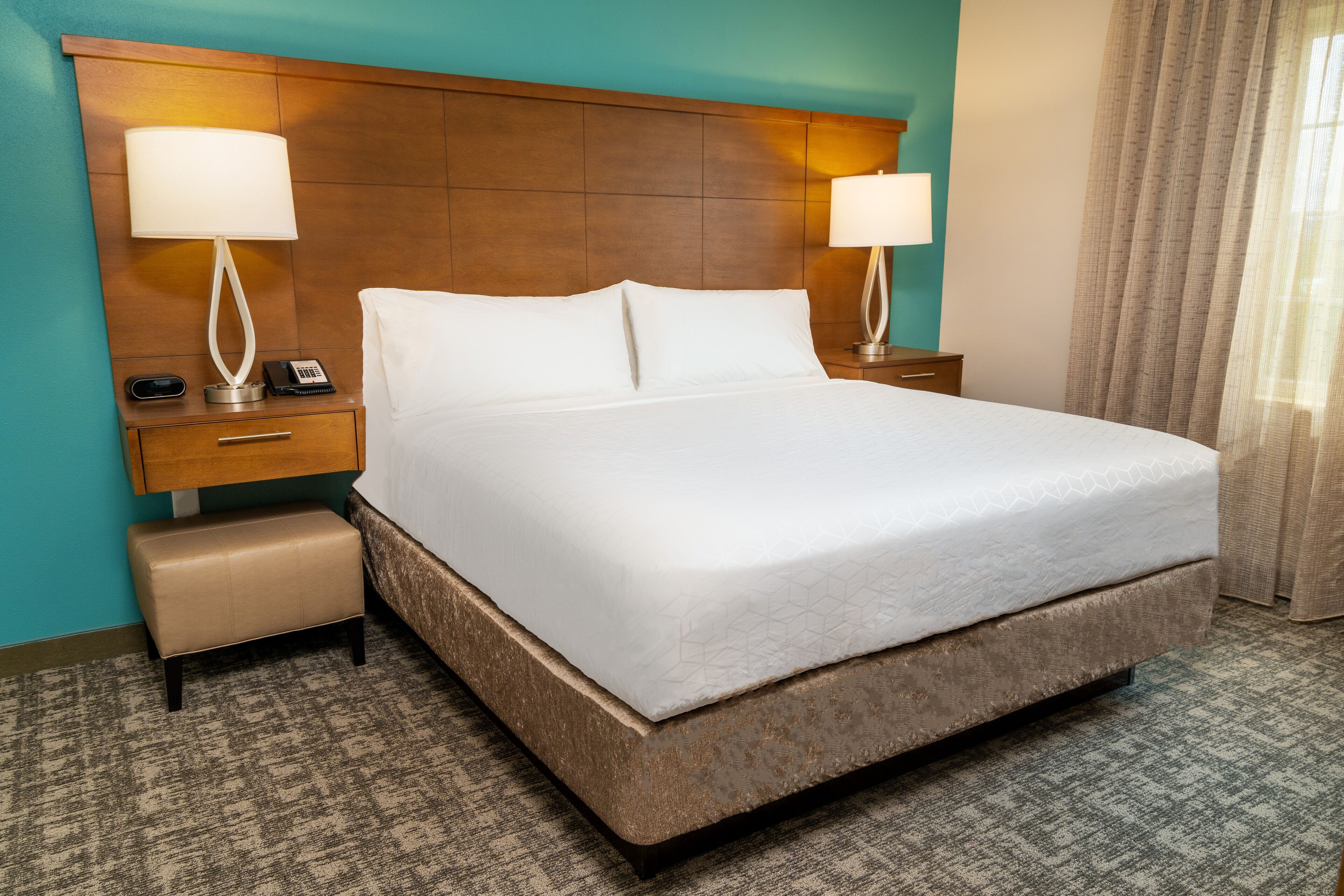 A hotel room with a large bed, two bedside tables with lamps, a small bench, and a wooden headboard.