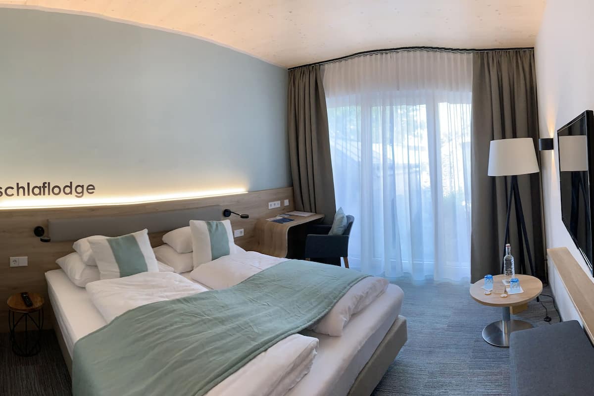 A hotel room with a bed, bedside table, lamp, TV, and a desk with a chair.