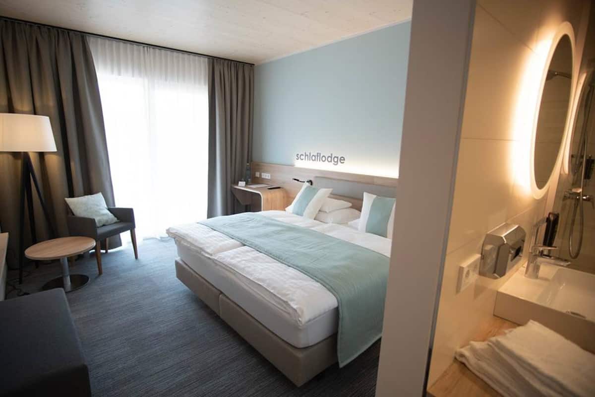 A hotel room with a bed, a desk, a chair, a lamp, a mirror, and a shower area.