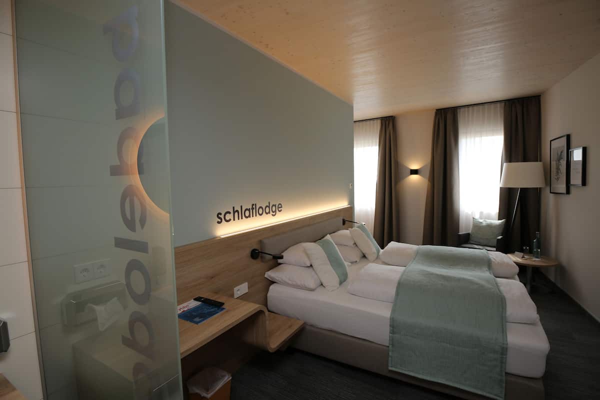 A hotel room with a bed, bedside table, lamp, and a wall with the hotel name.