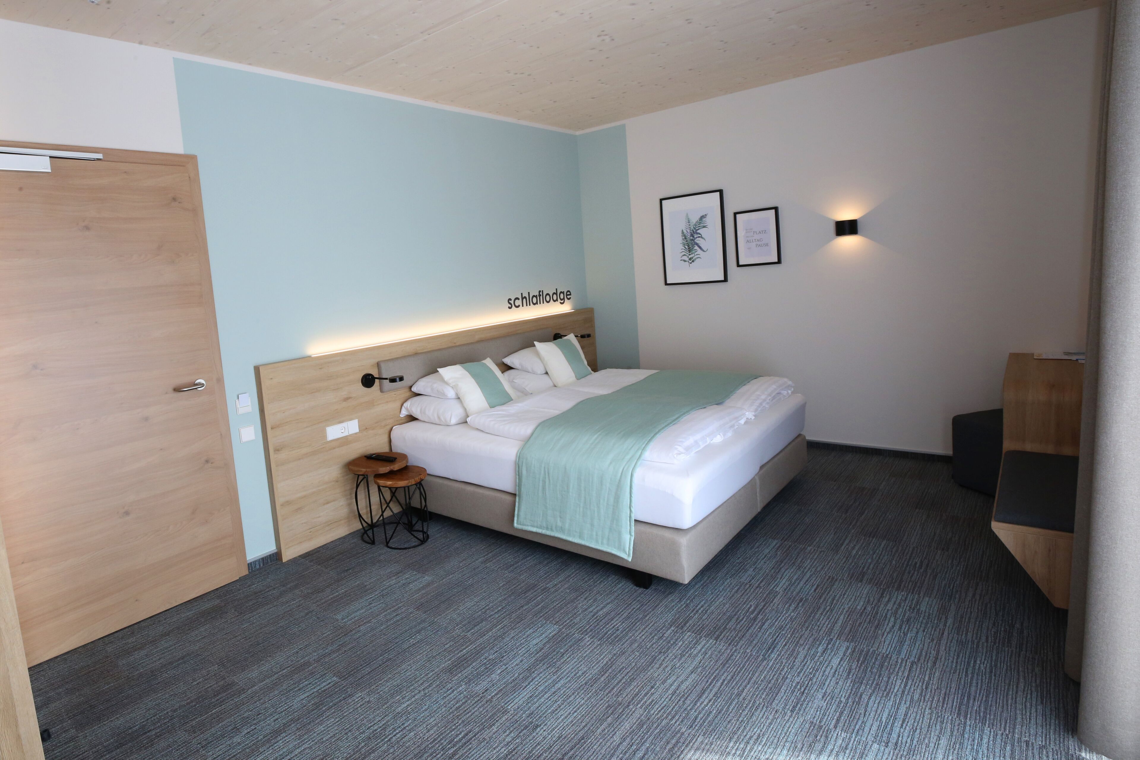 Une chambre à coucher comprenant un lit, une table de chevet, un petit tabouret, une porte en bois et un luminaire fixé au mur.