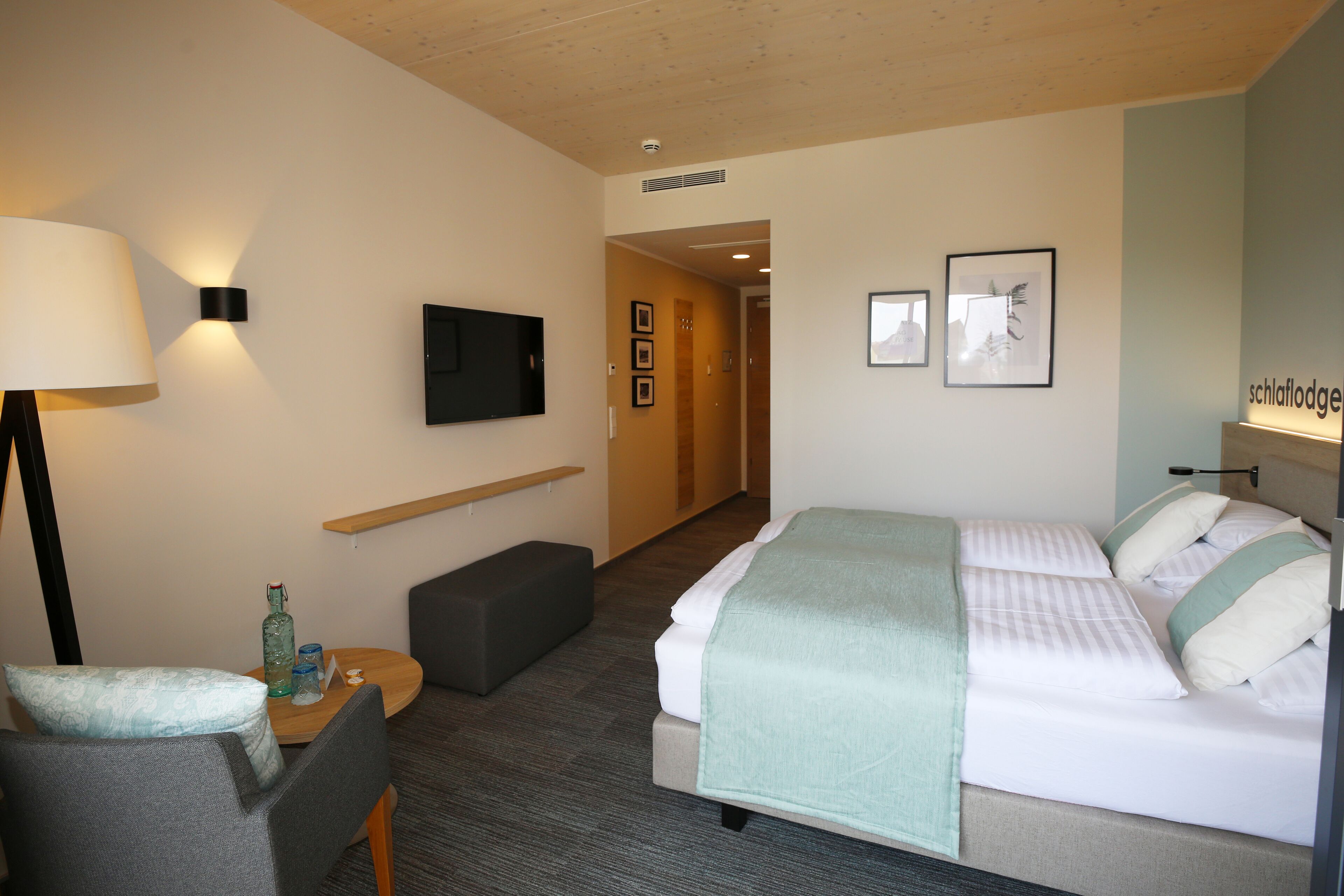A hotel room with a bed, a desk with a lamp, a chair, a small table with a bottle, and a television on the wall.