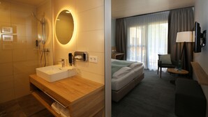 A hotel room with a bathroom featuring a sink, mirror, and shower. The room has a bed, a chair, a table, and a TV.