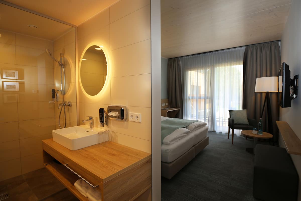 A hotel room with a bathroom featuring a sink, mirror, and shower. The room has a bed, a chair, a table, and a TV.