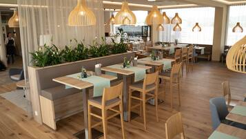 A modern restaurant with wooden tables and chairs, a central seating area with plants, and pendant lighting.