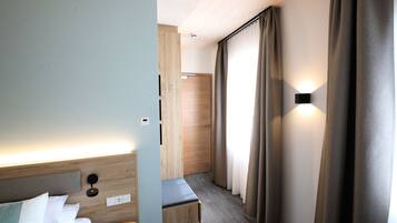 A hotel room with a bed, a bench, a wooden wardrobe, a door, and a window with curtains.