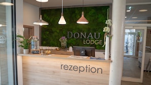 A reception desk with a wooden finish and the word "reception" on it. Behind the desk, there is a wall with a green leafy backdrop and the name "DonaU Lodge" displayed.