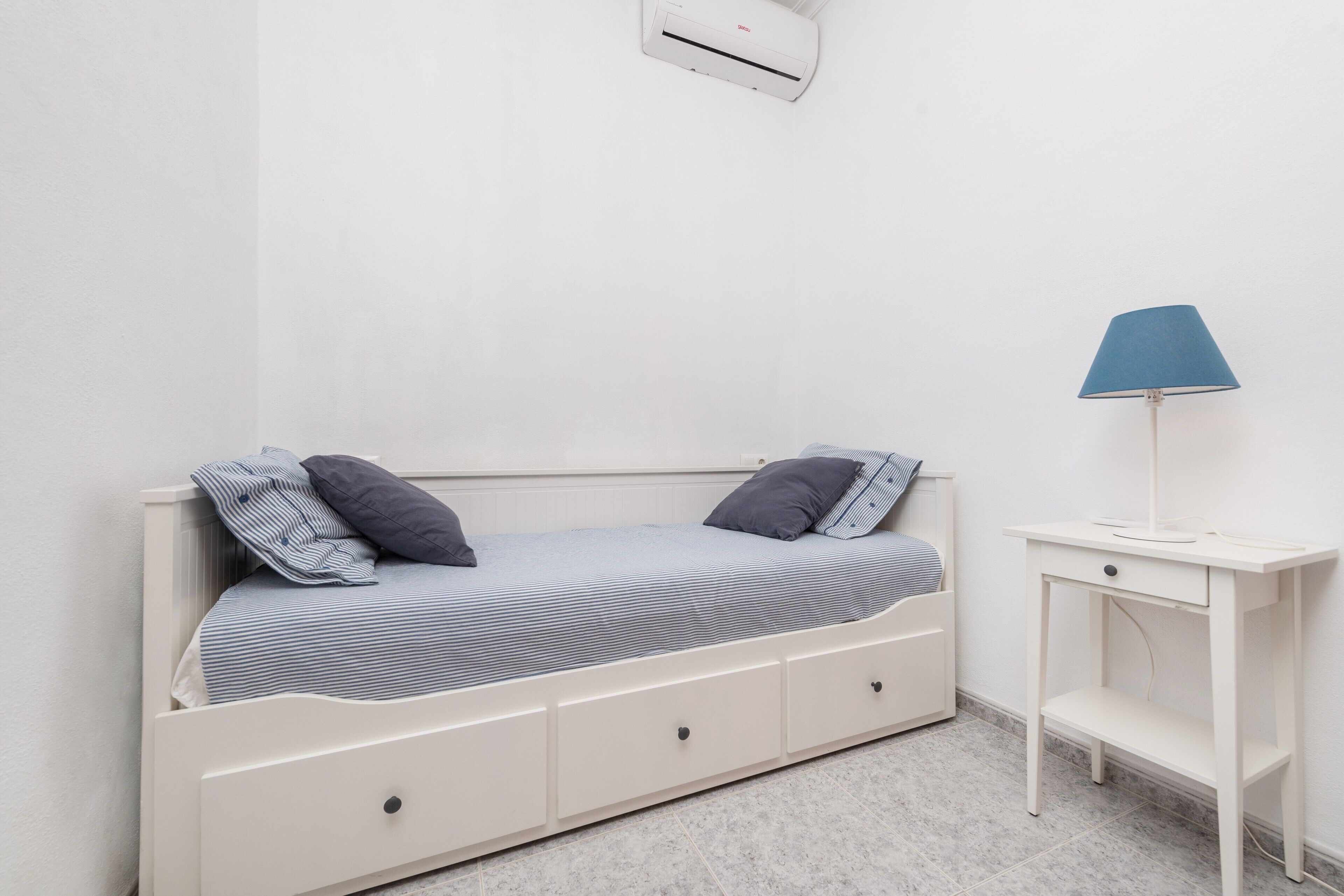 Habitación individual con una cama blanca, dos almohadas, una mesita de noche con lámpara y aire acondicionado instalado en la pared.