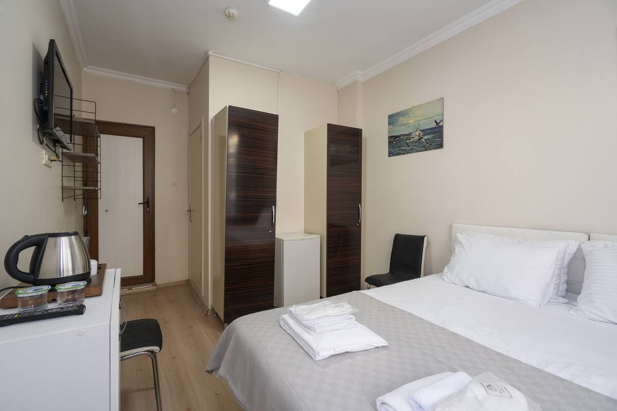A hotel room with a bed, a desk with a kettle, a chair, and a TV mounted on the wall.