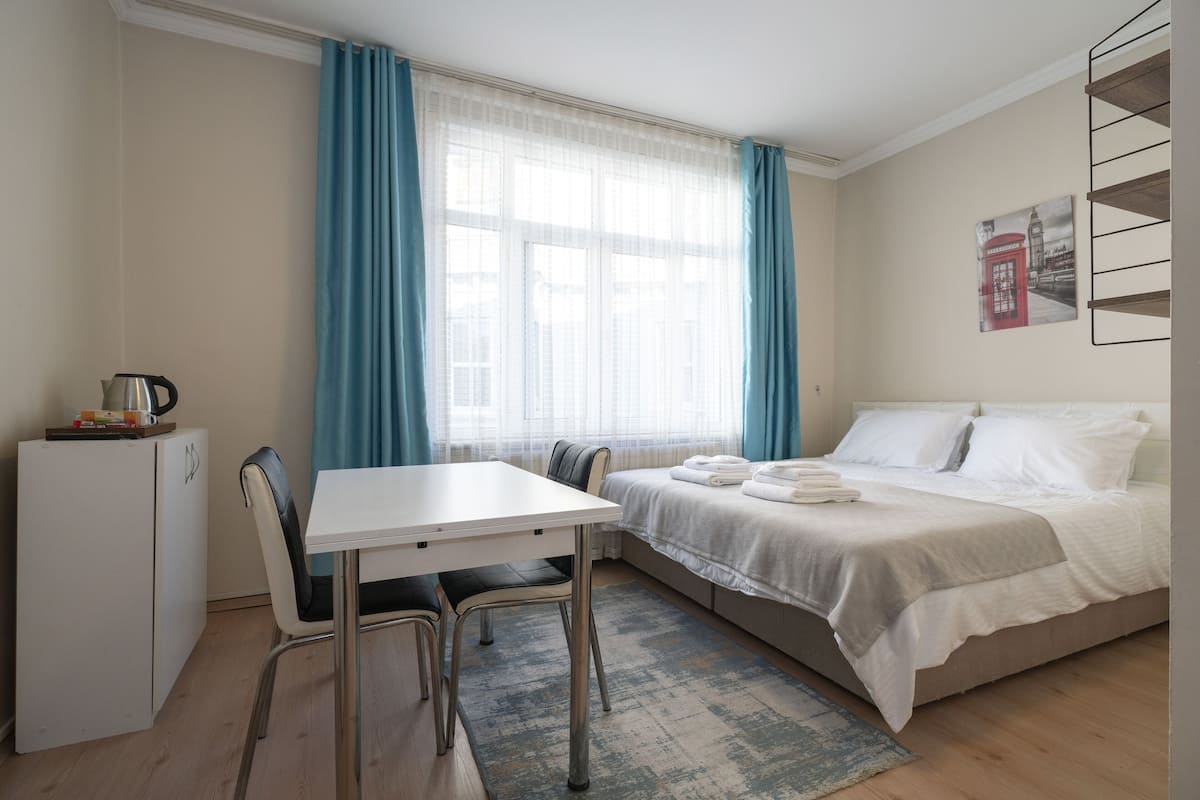 A compact hotel room with a bed, a small desk with a chair, a kitchenette, and a window with curtains.