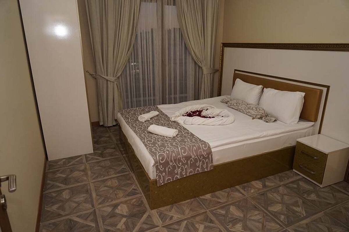 A neatly made bed with a patterned bedspread and pillows, a bedside table with a drawer, and a window with curtains.