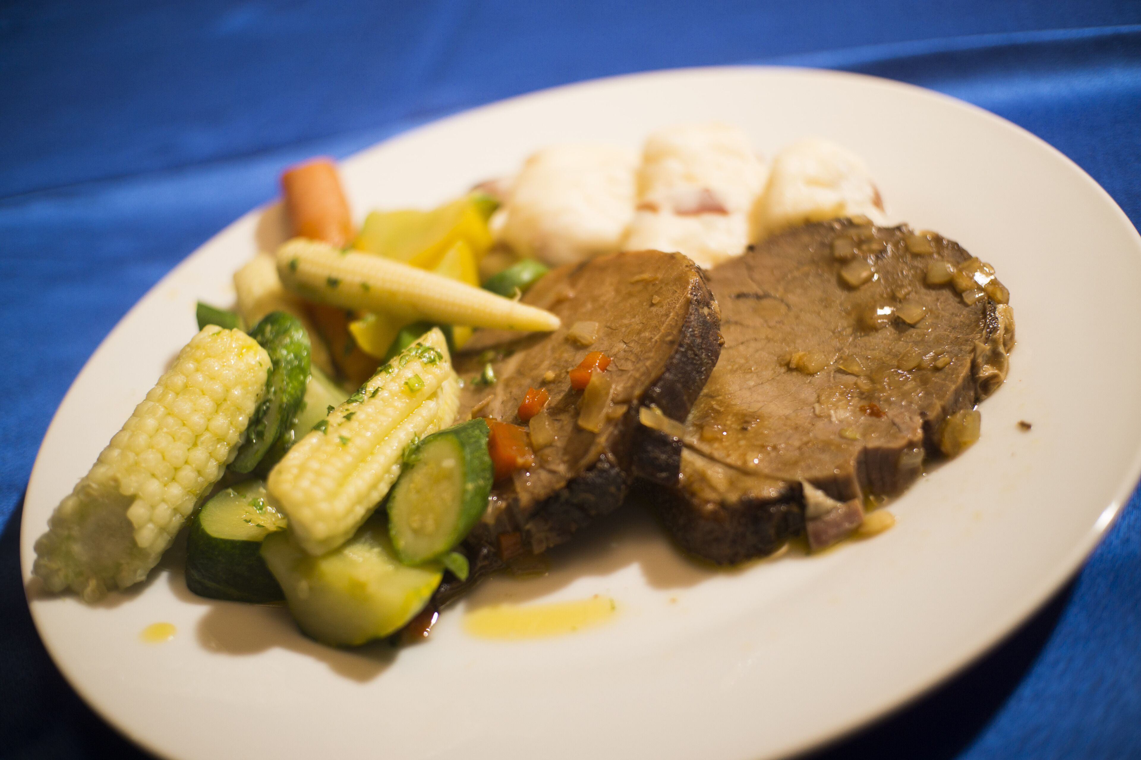 Une assiette remplie de nourriture, comprenant de la viande, des légumes et un aliment de couleur claire, peut-être du poisson, le tout posé sur une nappe bleue.