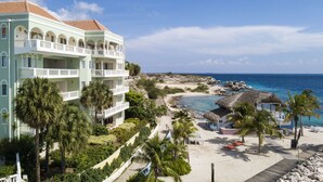 Resort com vista para a praia, incluindo um edifício com várias varandas, uma área de praia arenosa com palmeiras e uma estrutura com telhado de palha.