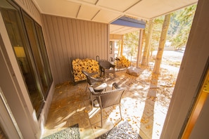 Un espace extérieur abrité avec des chaises en osier et une table, une pile de bois de chauffage et un sol carrelé parsemé de feuilles.