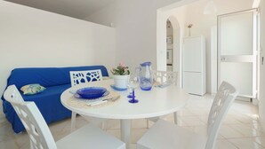 Une pièce moderne et bien éclairée, avec une table ronde blanche dressée pour le repas, un canapé bleu agrémenté de coussins décoratifs et une porte blanche donnant sur un autre espace.