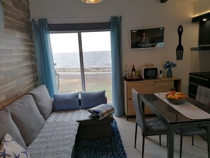 Ein modernes Wohnzimmer mit einer Couch, einem Esstisch mit Stühlen, einer Küchenzeile und einem großen Fenster mit Meerblick.