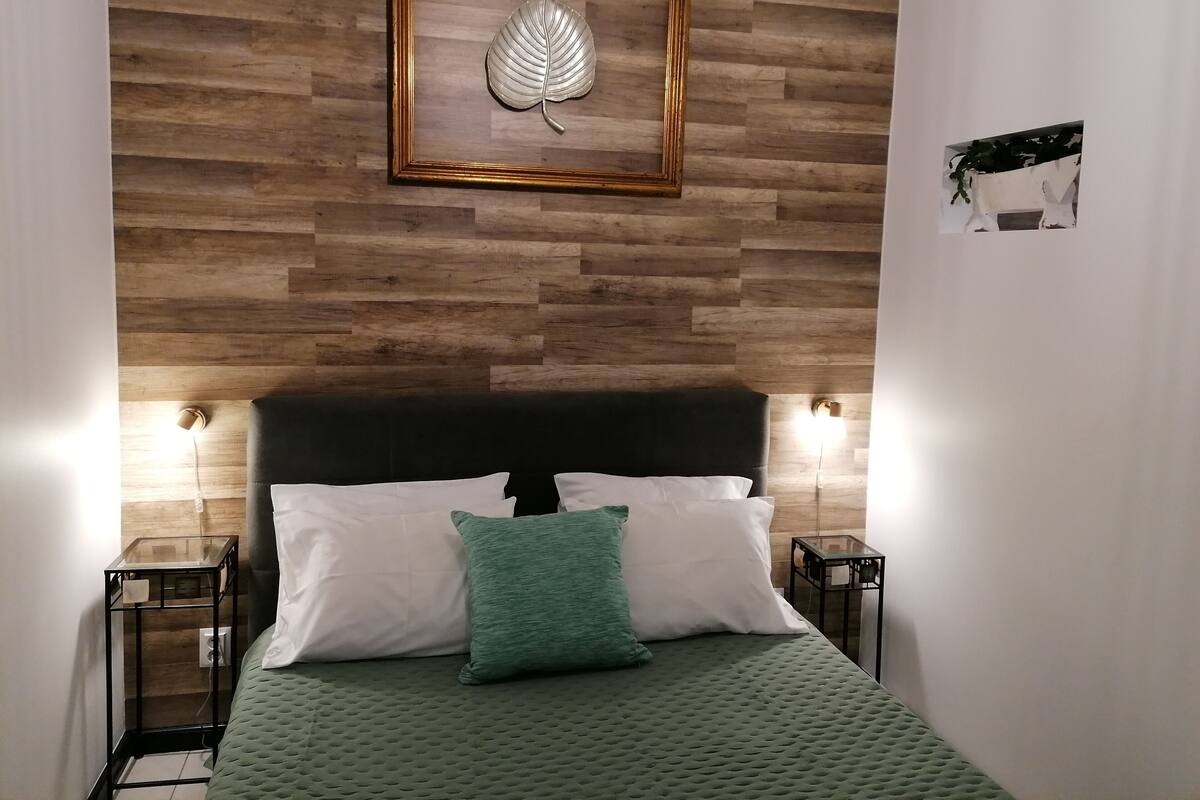 Ein ordentlich eingerichtetes Schlafzimmer mit Holzvertäfelung, einer gerahmten Blattform-Kunstwerk, einer grünen Bettdecke und zwei Nachttischen mit Lampen.