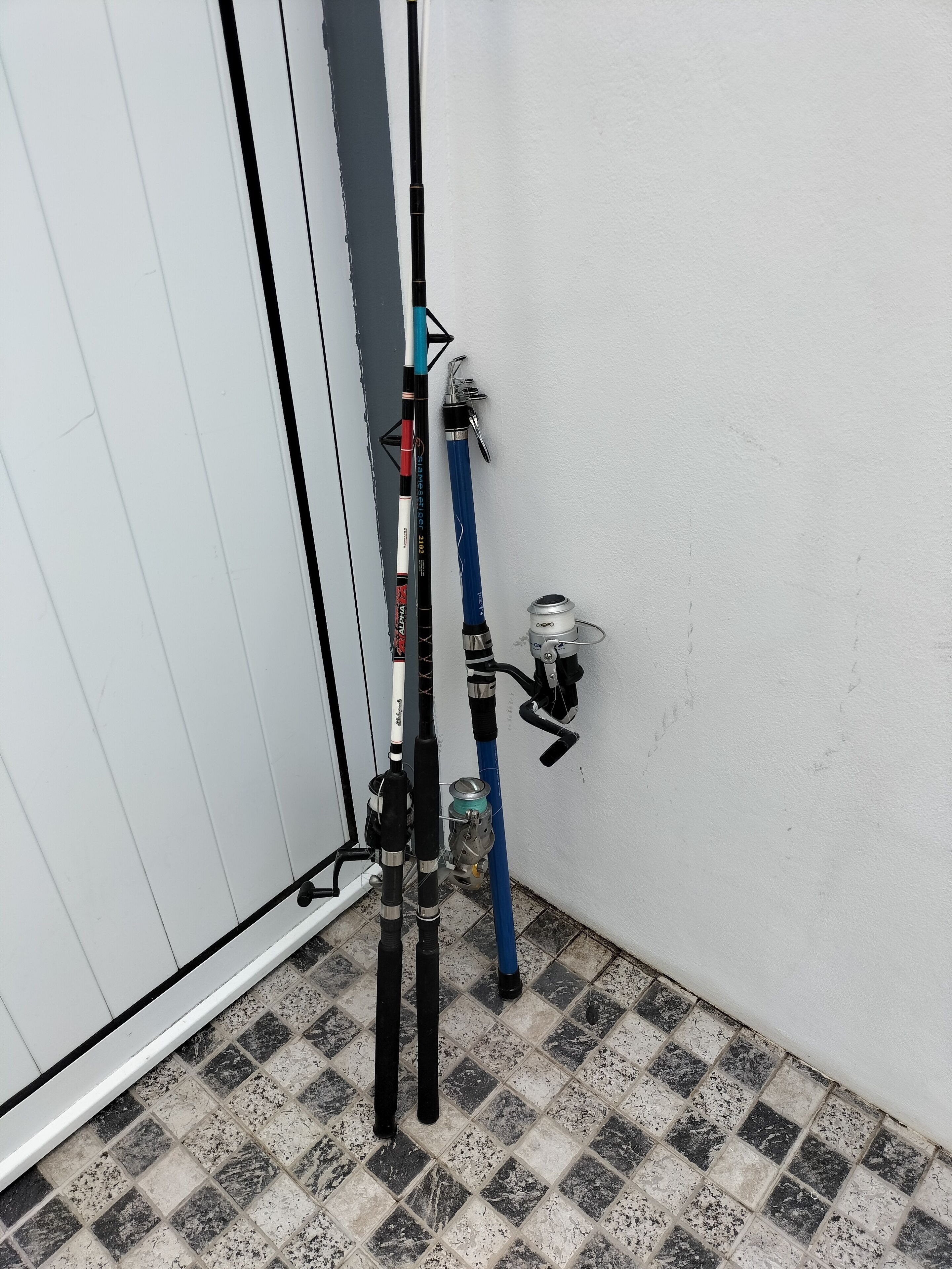 Canto de um quarto com piso de azulejos e dois equipamentos de pesca encostados em uma parede branca.