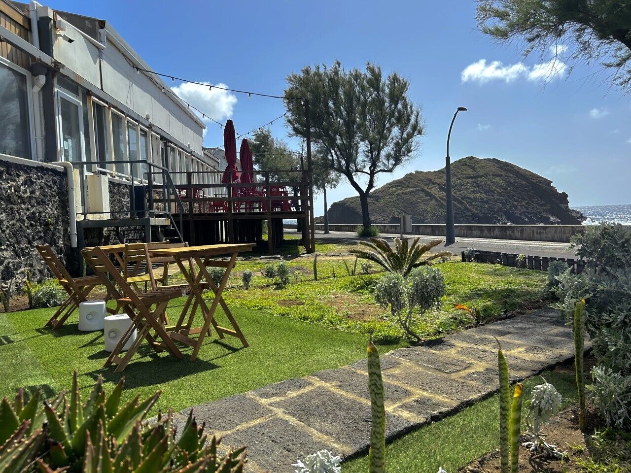 A patio area with a wooden table and chairs, a small garden with various plants, and a view of the sea and a hill in the background.