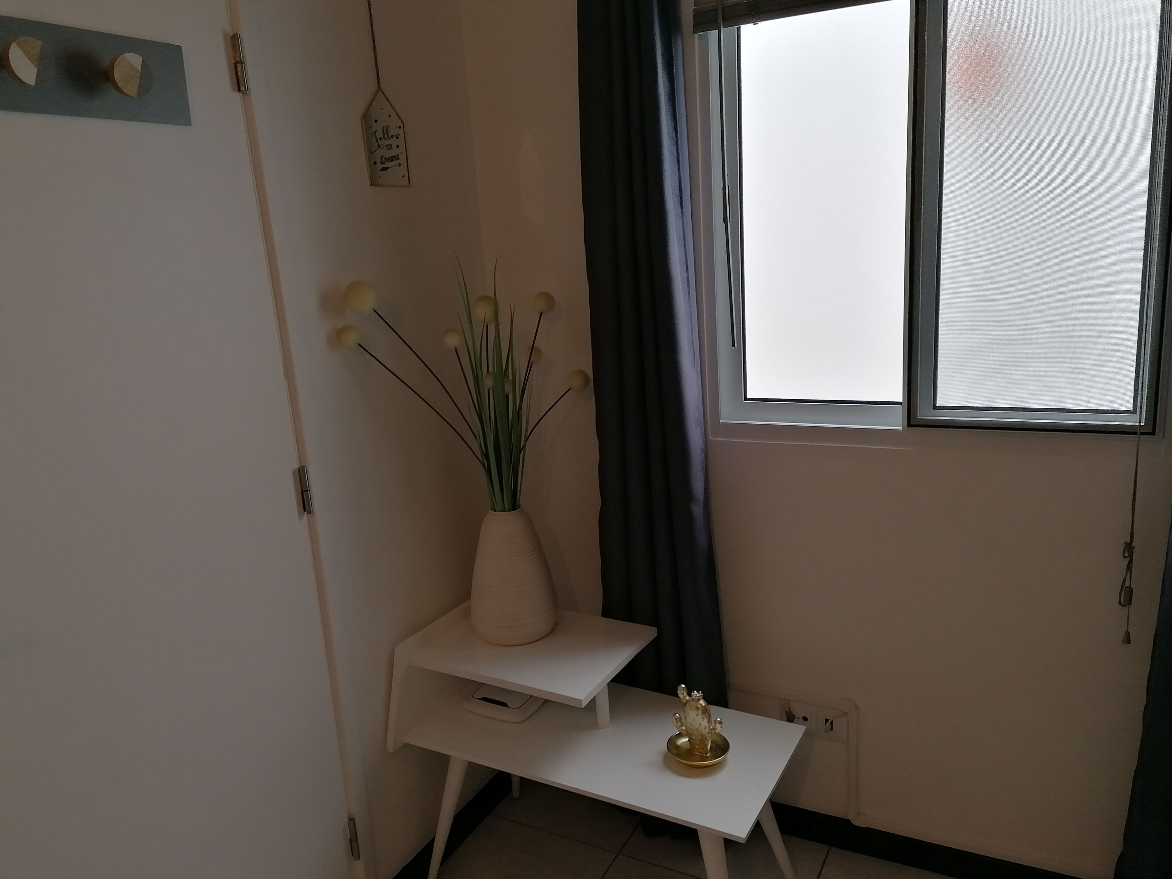 Une pièce avec un bureau blanc, un vase contenant des plantes vertes, un petit cactus et une fenêtre avec des rideaux.