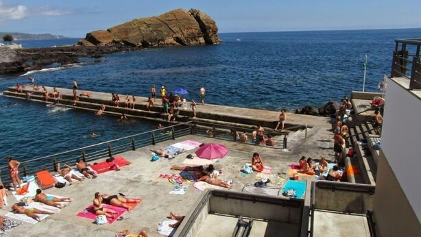 Een kustgebied met een rotsachtige uitstulping, een zwemgedeelte en mensen die op een terras zonnebaden.