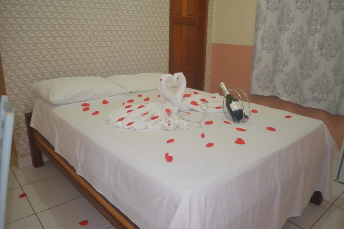 A neatly made bed with a white bedspread, a heart-shaped towel arrangement, and scattered rose petals.